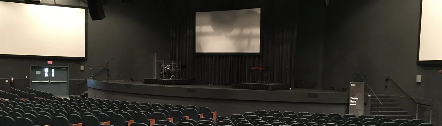 An empty auditorium with rows of seats facing a stage, projection screens and a drum kit.