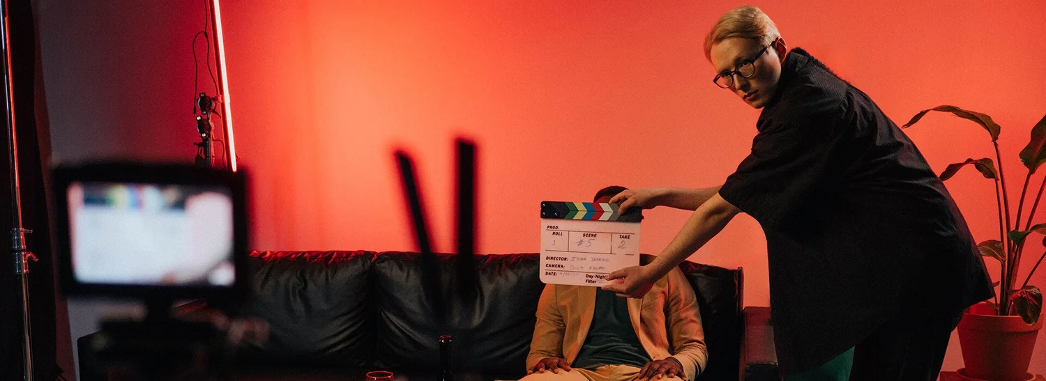 A crew member holds a clapperboard in front of a seated contributor on a red-lit set.