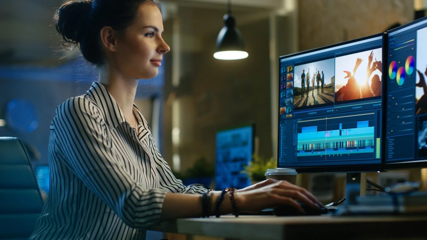 A video editor working at a desk with two monitors showing a timeline and colour tools.