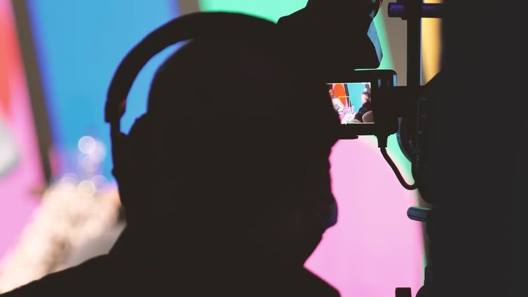 Silhouette of a camera operator in headphones filming a live event, the camera monitor glowing against bright pink and teal stage graphics.