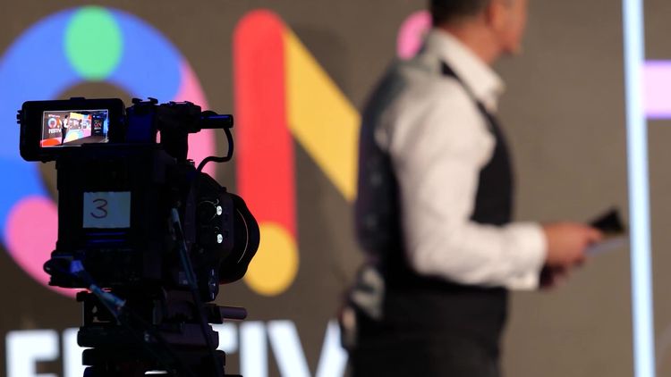 A broadcast camera on a tripod filming a presenter on stage in front of a large, colourful festival backdrop.