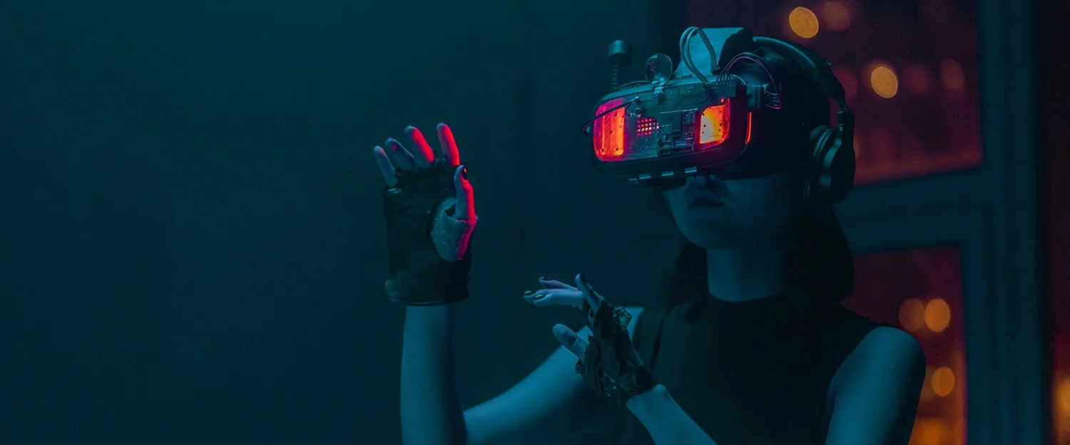 A woman in a glowing VR headset and haptic gloves reaching out in a dark room.