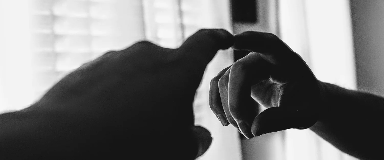Black-and-white silhouette of two hands about to touch fingertips against a bright window.