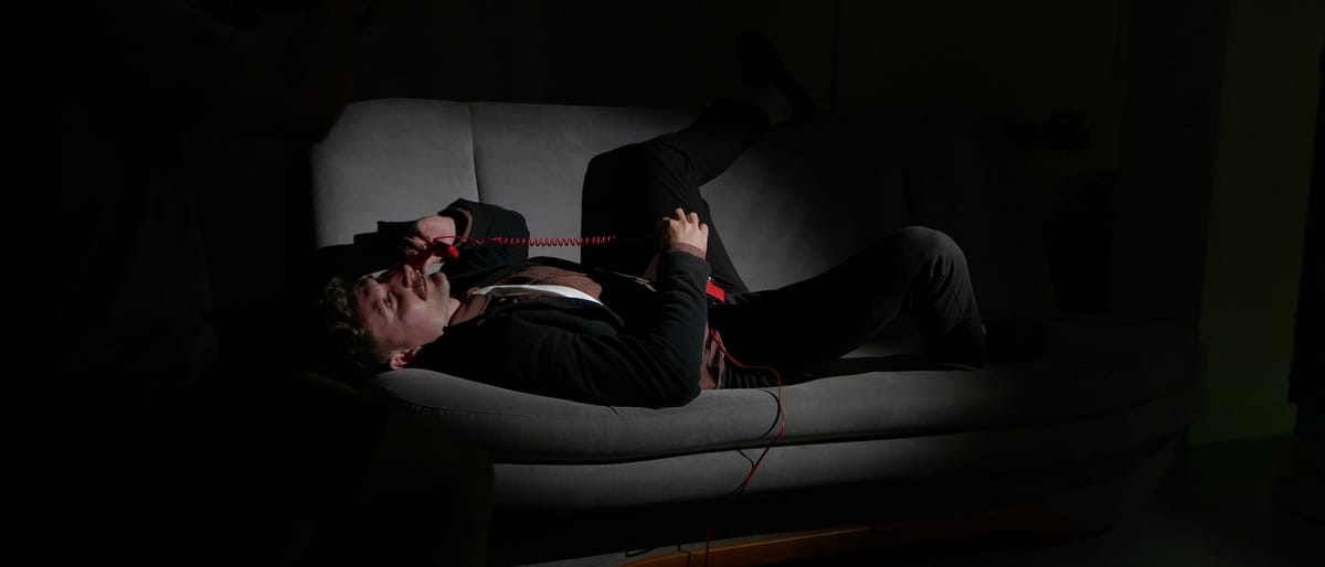 A man lies on a sofa in a dark room holding a red telephone, a still from the Strange In Stereo music video When You Say My Name, where Nigel assisted on camera.