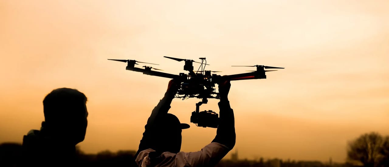 Silhouette of a person launching a camera drone against an orange sunset sky.