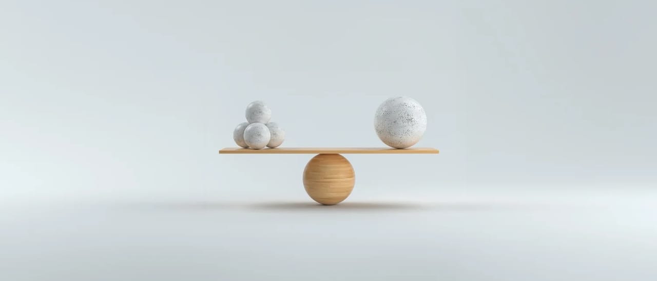 Two spheres balanced on a simple seesaw on a pale grey background.