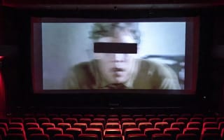 A cinema screen showing a face with a black censor bar across the eyes, above rows of red seats.