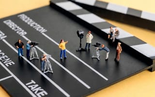 Miniature film-crew figures standing on a full-size clapperboard against a yellow backdrop.