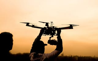 Silhouette of a person launching a camera drone against an orange sunset sky.